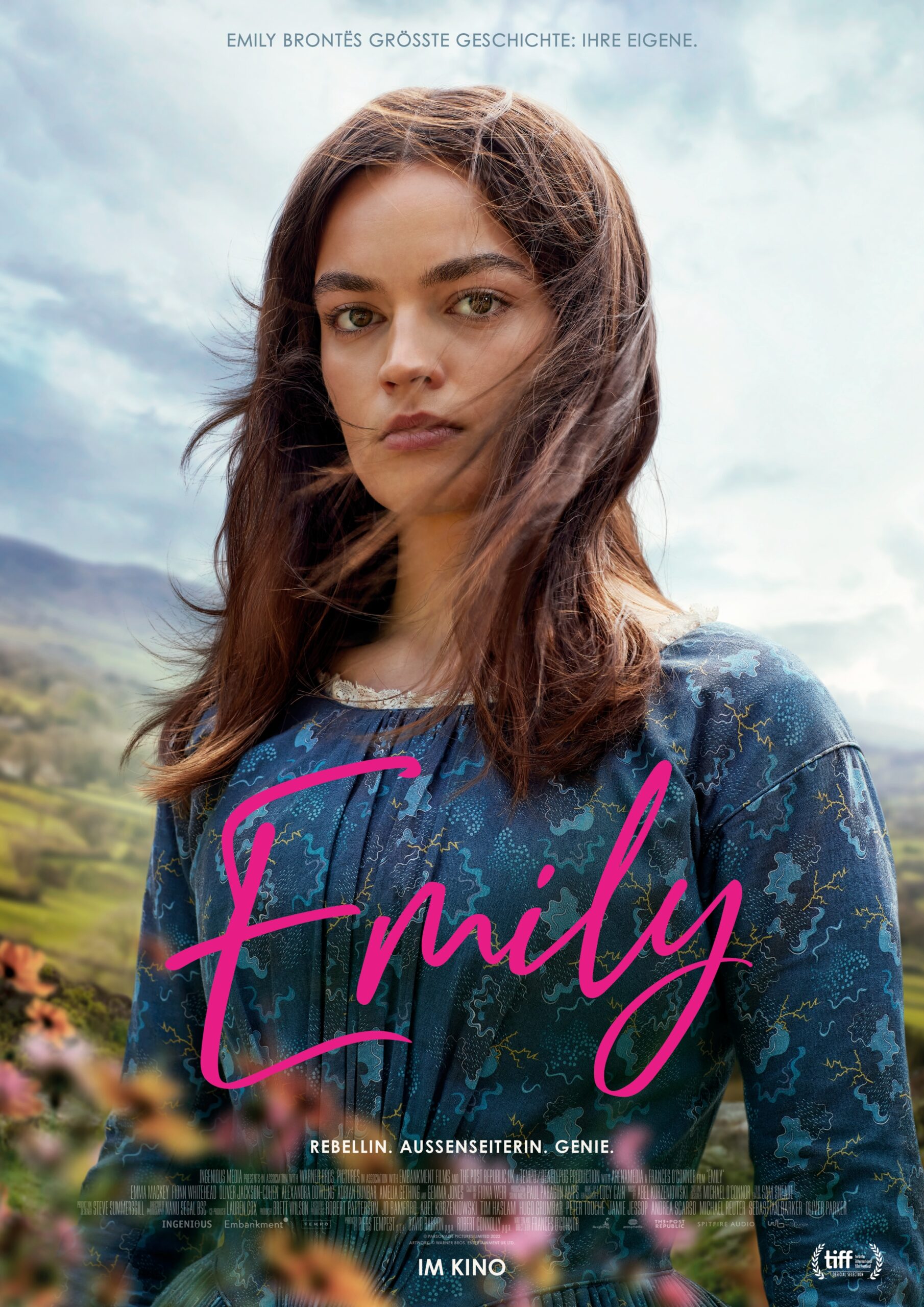 Filmplakat zu EMILY: Emma Mackey als Emily Brontë im Bruststück, vor einer Hügellandschaft mit wolkigem Himmel, über ihrem blauen Kleid eine Handschrift in Magenta "Emily"
